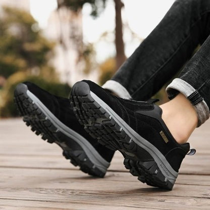 Black slip-on trail shoe worn on foot walking on wooden planks