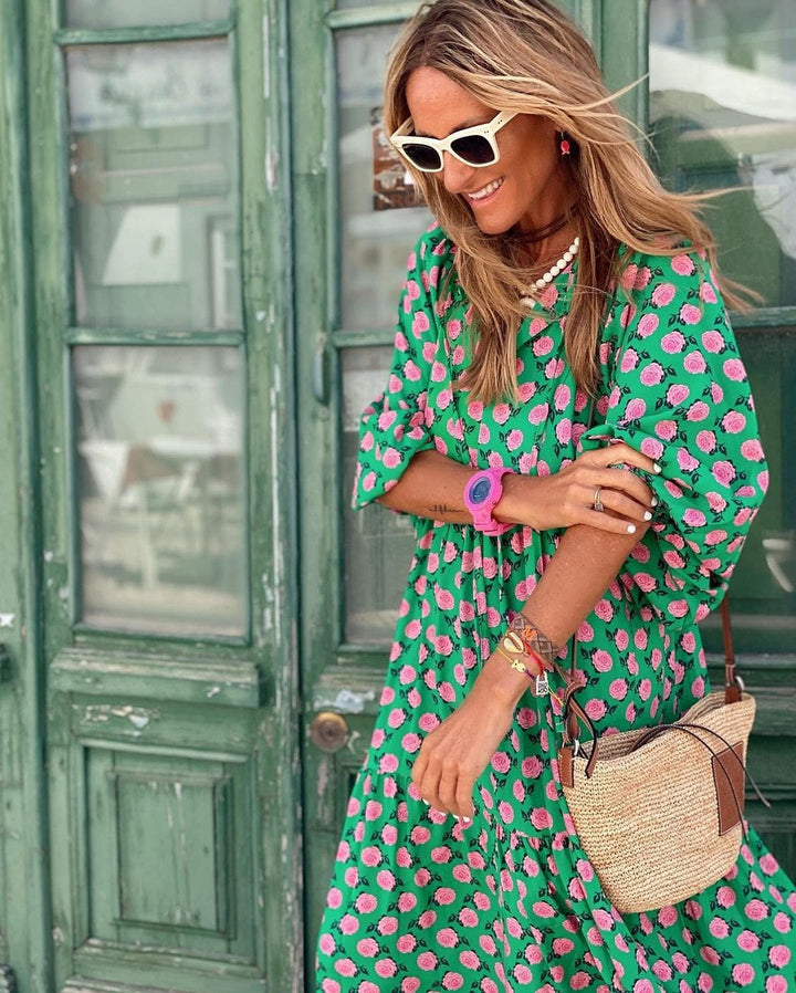 Model in green floral maxi dress adjusting pink watch, straw bag in hand