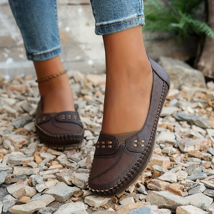 brown slip-on loafers with stitched trim shown mid-step on gravel