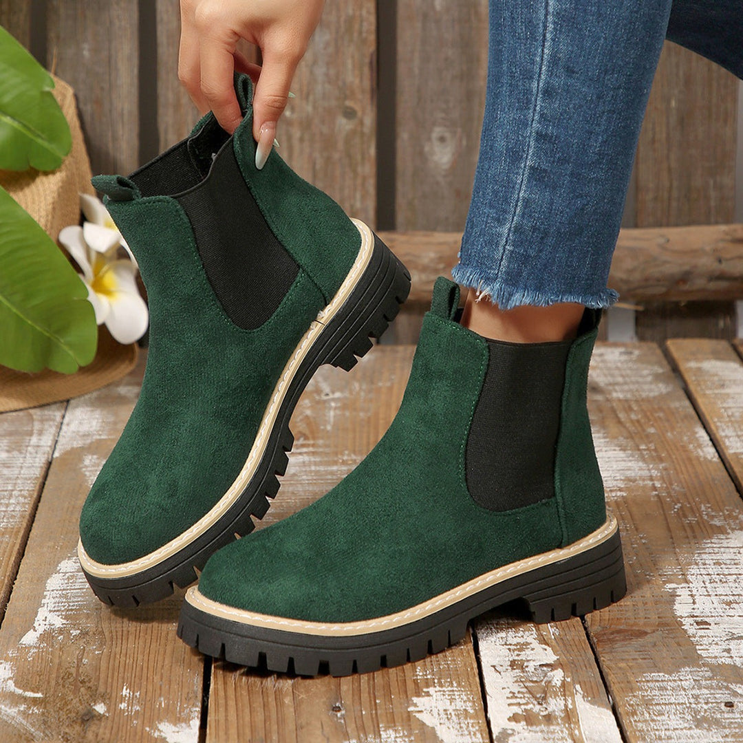 Dark green suede Chelsea boots being lifted by hand on wooden floor