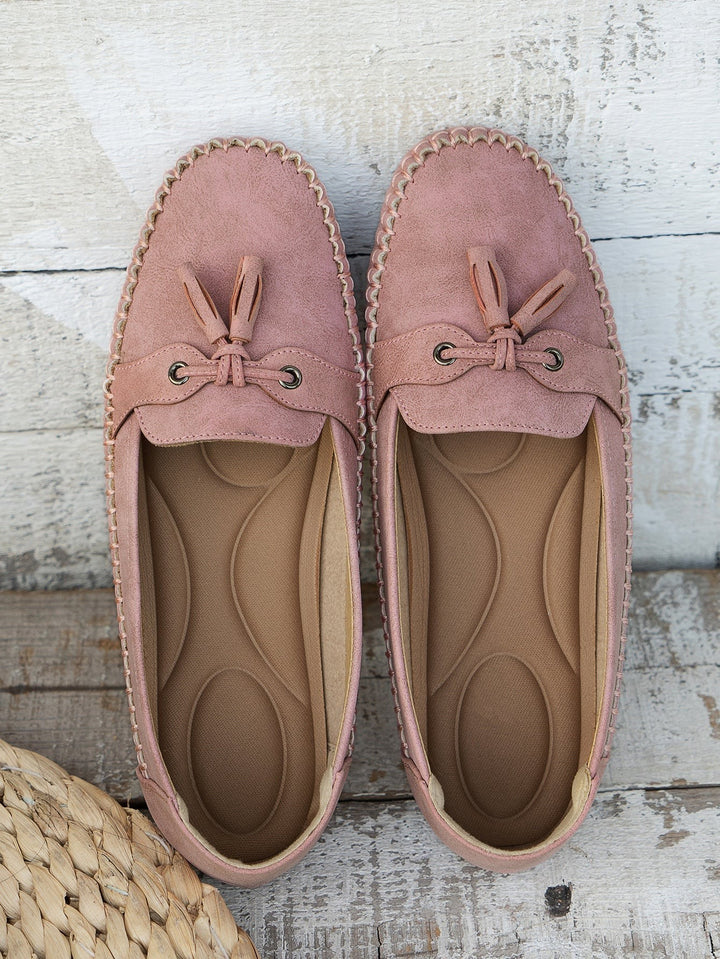Dusty pink suede loafers with tassel bow displayed top-down on wood floor