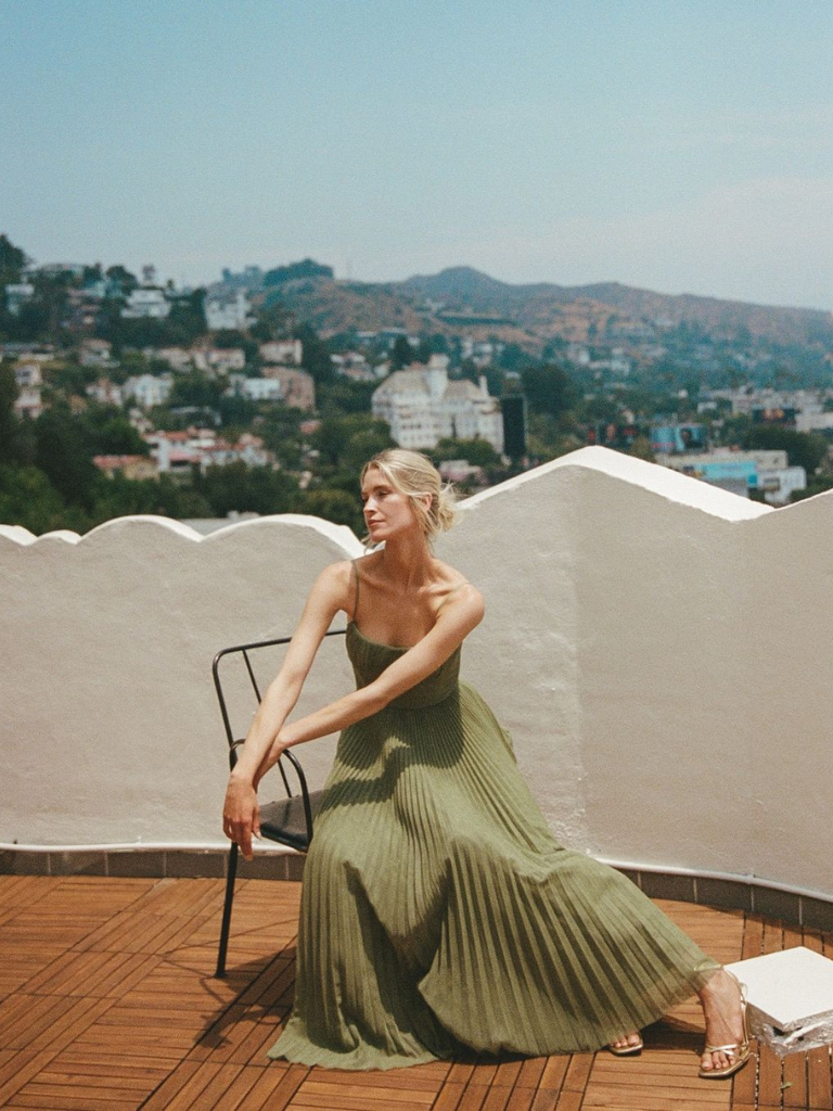 Woman in olive pleated maxi dress seated outdoors on rooftop terrace
