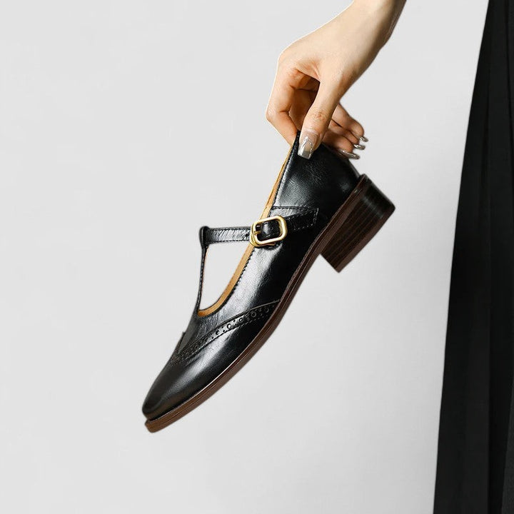 Black leather T-strap Mary Jane shoe dangling from fingertips, side view