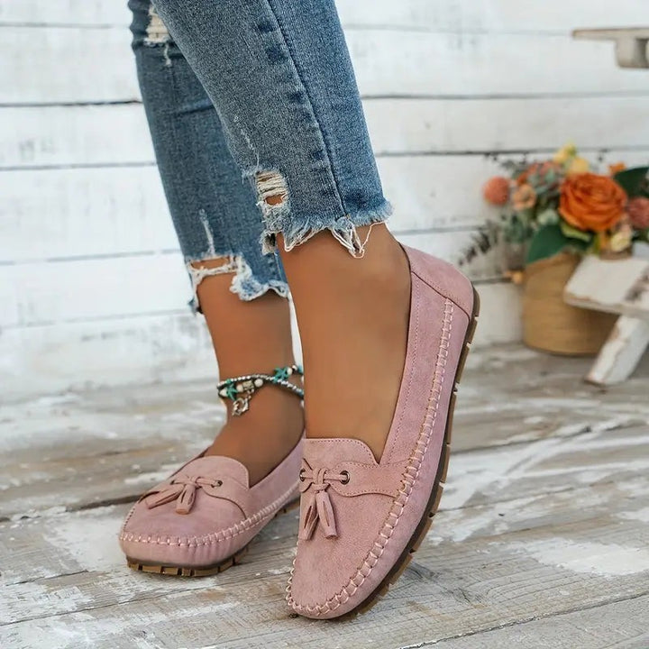 Pink suede moccasin loafers worn barefoot against rustic wood backdrop