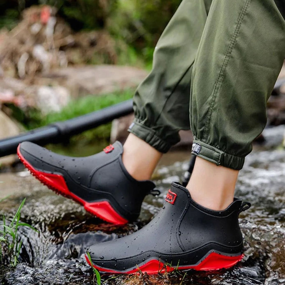 Black waterproof ankle boots with red sole, worn beside a stream