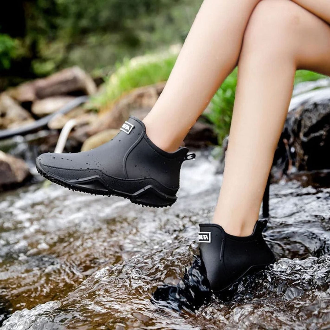 Black slip-on rain boots with white sole detail, worn near a stream